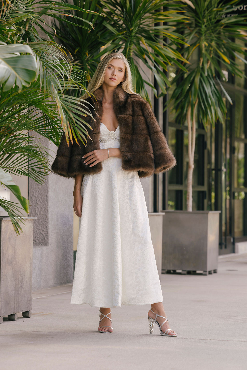 Woman in a white dress with a fur coat and silver heels standing in an outdoor setting with plants.