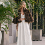 Woman in a white dress with a fur coat standing among tropical plants.