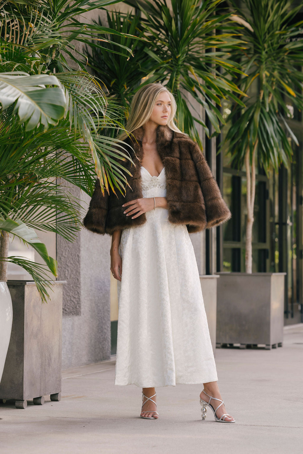 Woman in a white dress with a fur coat standing among tropical plants.
