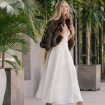 Woman in a white dress and brown fur coat standing among potted plants.