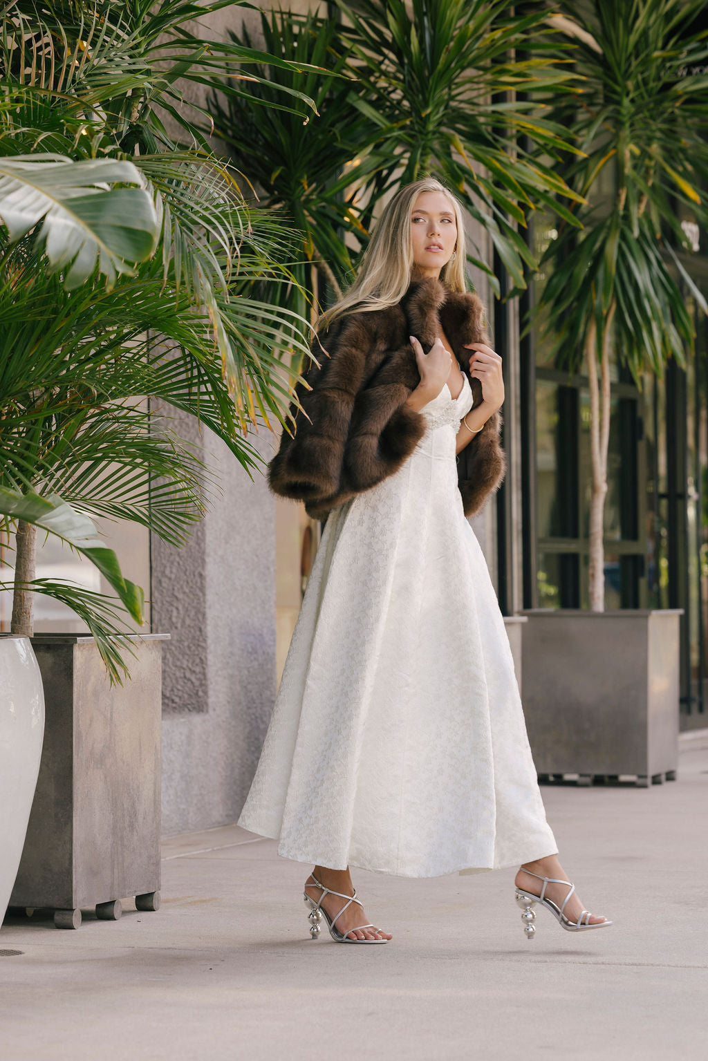 Woman in a white dress and brown fur coat standing among potted plants.