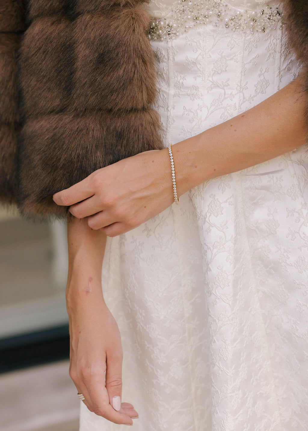 Close-up of a person wearing a white lace dress with a fur stole, focusing on the hand and bracelet.
