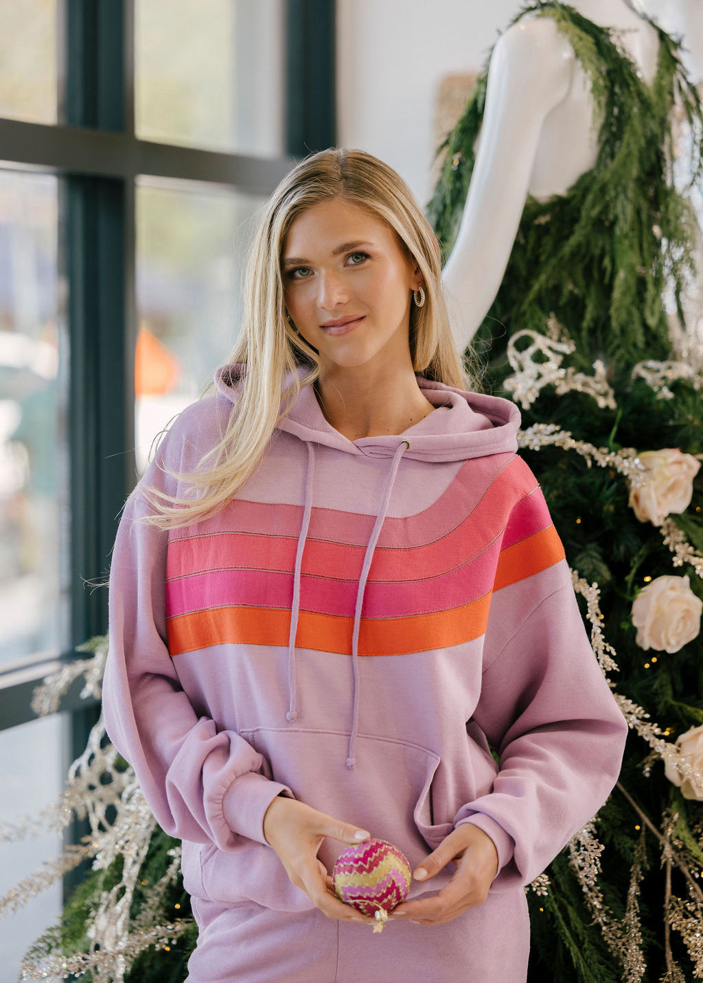Woman wearing a pink striped hoodie holding a cupcake in front of a decorated Christmas tree.