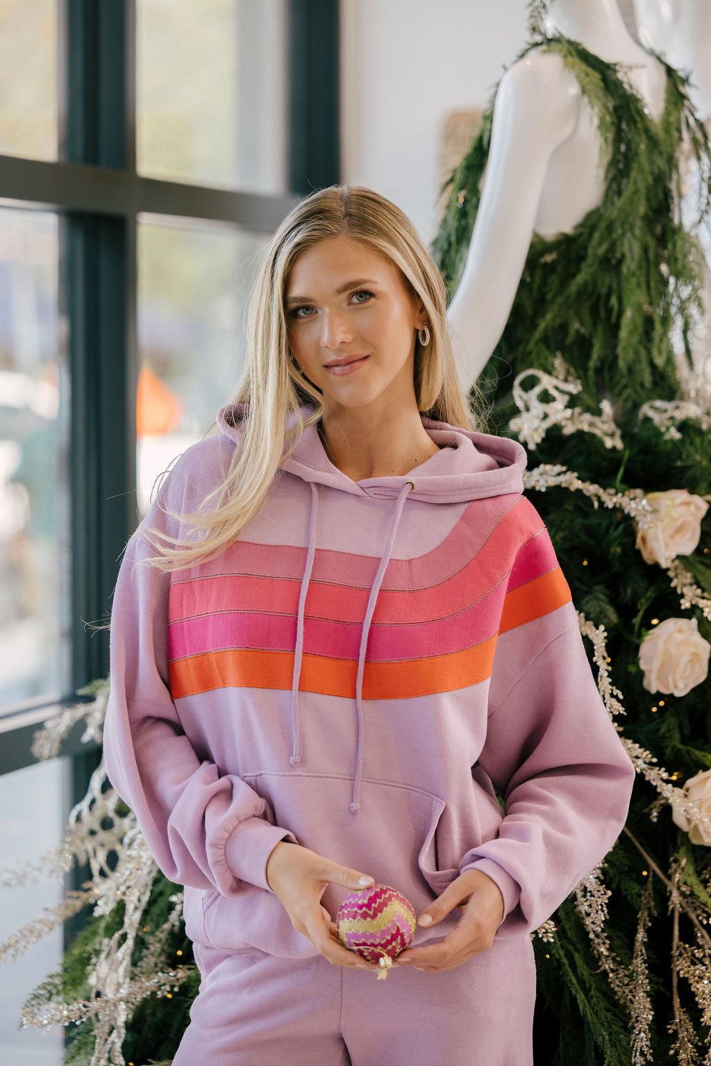 Woman wearing a pink striped hoodie holding a cupcake in front of a decorated Christmas tree.