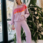 Woman wearing a mauve hoodie with a colorful stripe and matching pants, standing in front of a decorated Christmas tree.