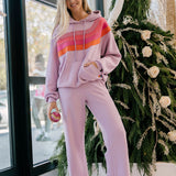 Woman wearing a mauve hoodie with a colorful stripe and matching pants, standing in front of a decorated Christmas tree.
