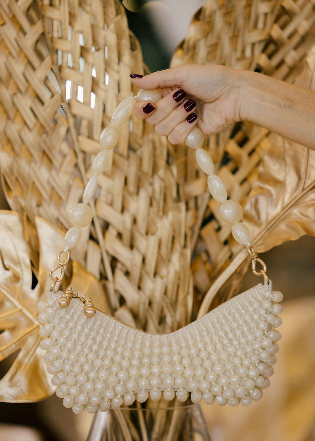 Hand holding a pearl-handled handbag against a textured wall with dried plants.