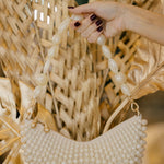 Hand holding a pearl-handled handbag against a textured wall with dried plants.
