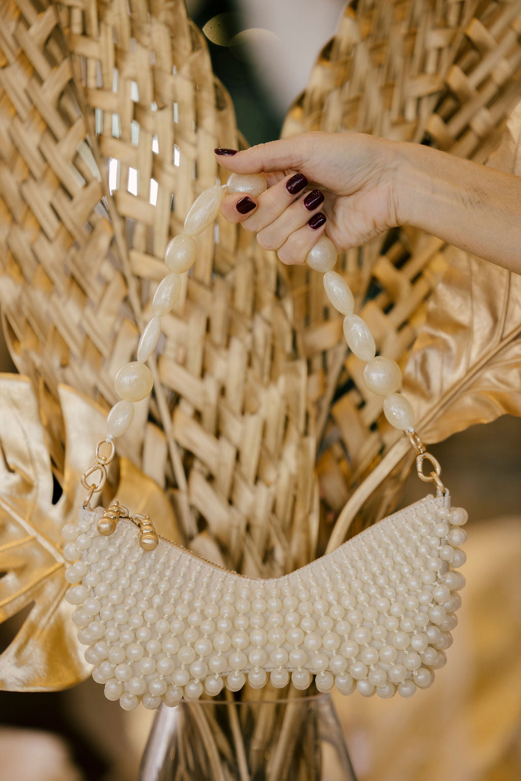 Hand holding a pearl-handled handbag against a textured wall with dried plants.