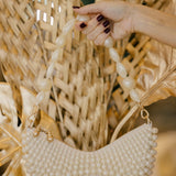 Hand holding a pearl-handled handbag against a textured wall with dried plants.