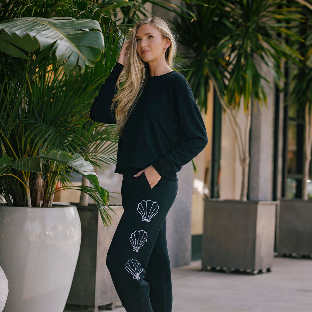 Woman wearing a black outfit with shell designs, standing in an outdoor setting with plants.