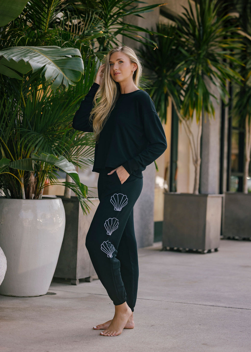 Woman wearing a black top and bottom with shell designs embroidered on pants, standing in an outdoor setting with plants.