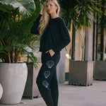 Woman wearing a black top and bottom with shell designs embroidered on pants, standing in an outdoor setting with plants.