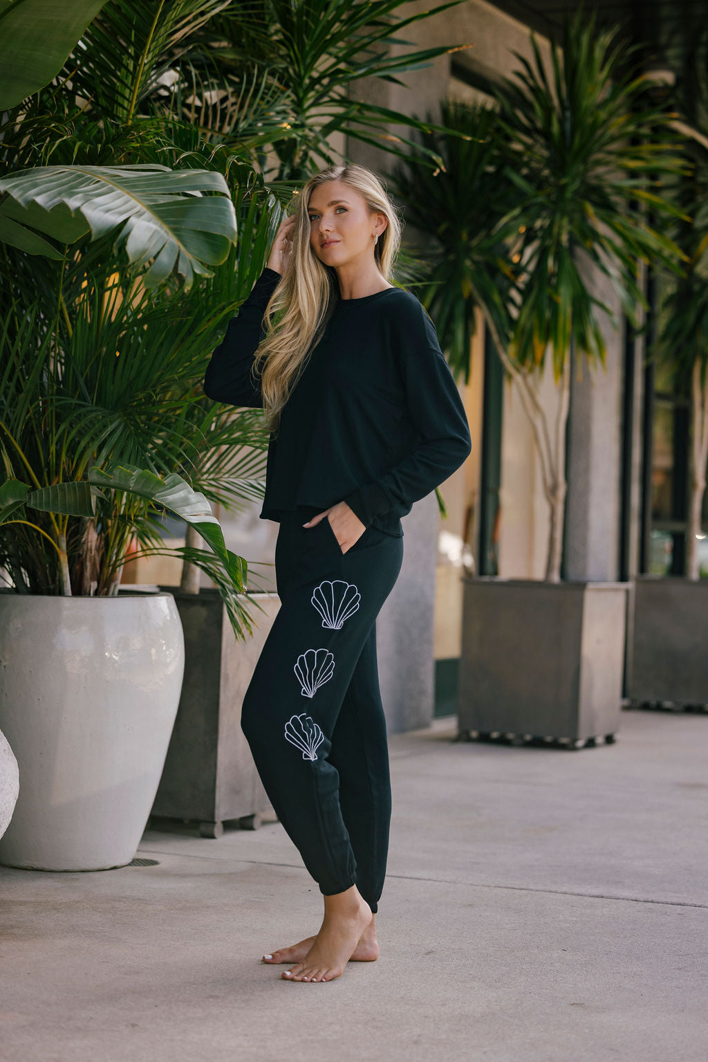 Woman wearing a black top and bottom with shell designs embroidered on pants, standing in an outdoor setting with plants.
