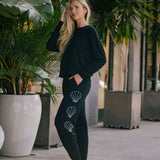 Woman wearing a black top and bottom with shell designs embroidered on pants, standing in an outdoor setting with plants.