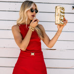 Woman in a red top and skort holding a gold clutch and wearing a gold bracelet against a white wooden background