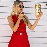 Woman in a red top and skort holding a gold clutch and wearing a gold bracelet against a white wooden background