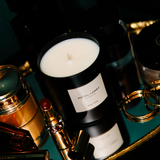 Candle in a black container with a label on a reflective surface, surrounded by decorative items.