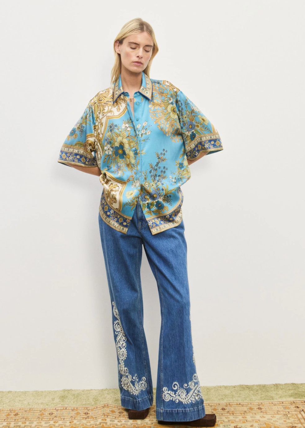 Person wearing a floral blouse and blue jeans with embroidery on a plain background