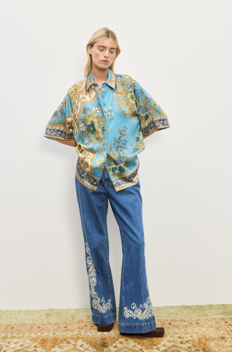 Person wearing a floral blouse and blue jeans with embroidery on a plain background
