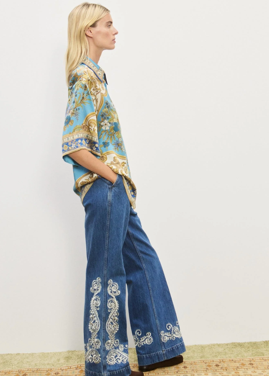 Woman wearing a patterned blouse and blue jeans with white embroidery on a plain background