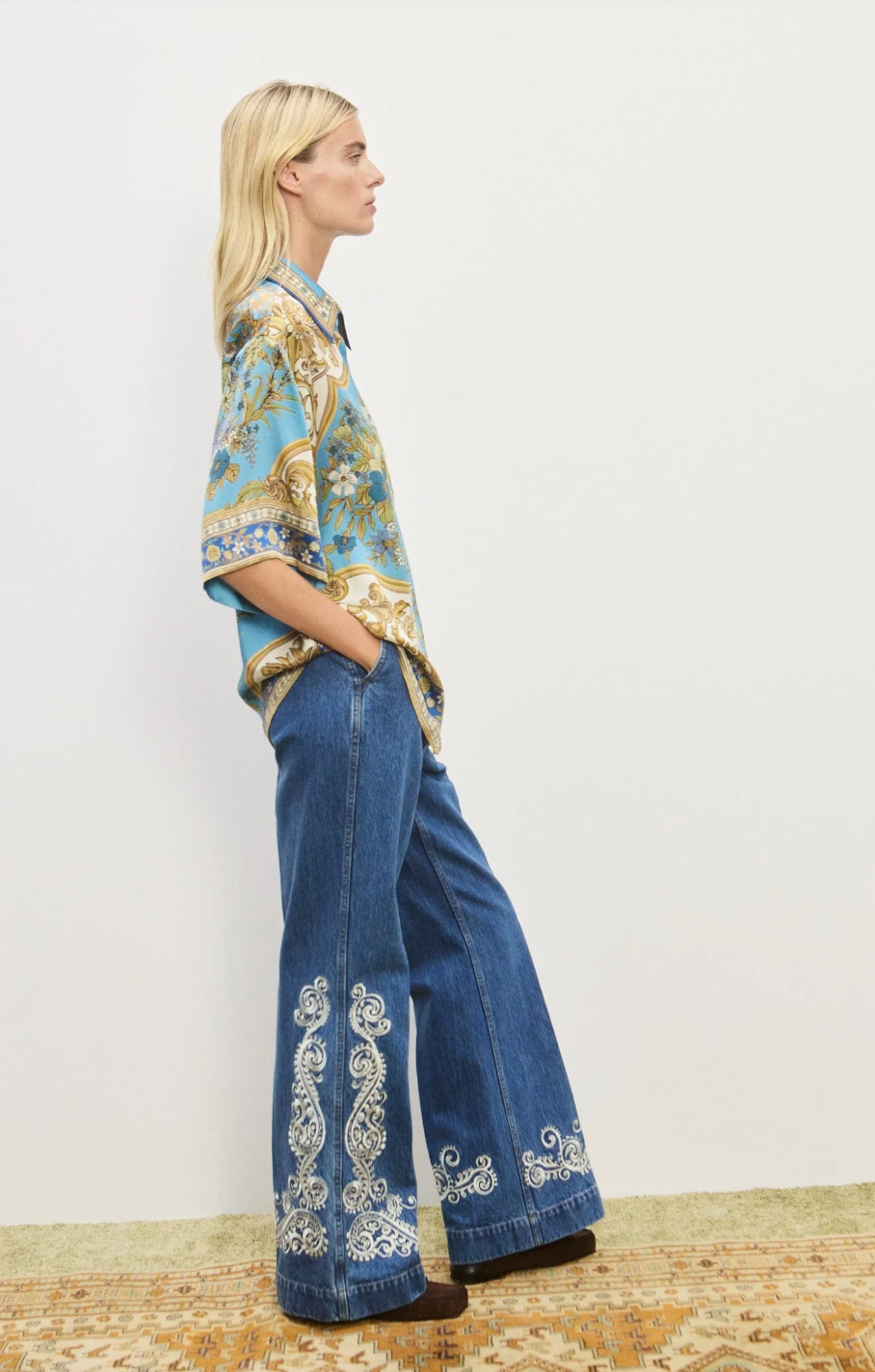 Woman wearing a patterned blouse and blue jeans with white embroidery on a plain background
