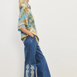 Woman wearing a patterned blouse and blue jeans with white embroidery on a plain background