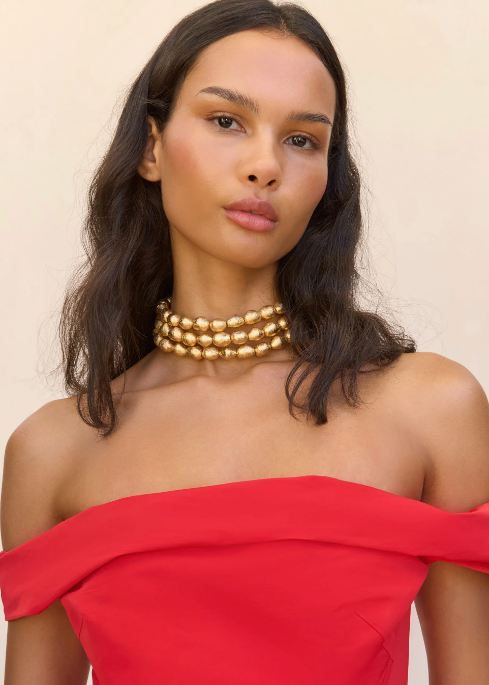 Woman wearing a gold necklace and red off-shoulder top against a beige background