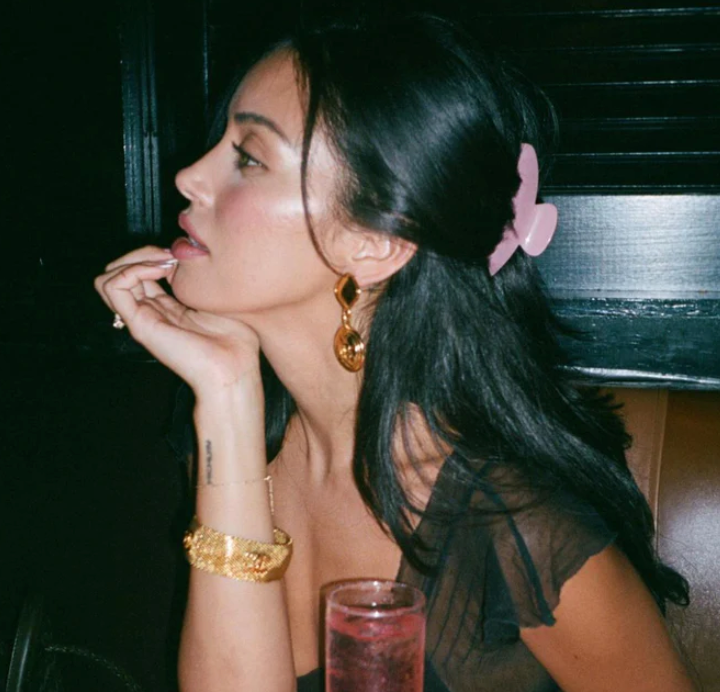 Woman with a pink hair clip sitting indoors, wearing gold jewelry.
