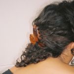 Person with curly hair wearing a wooden hair clip against a white wall