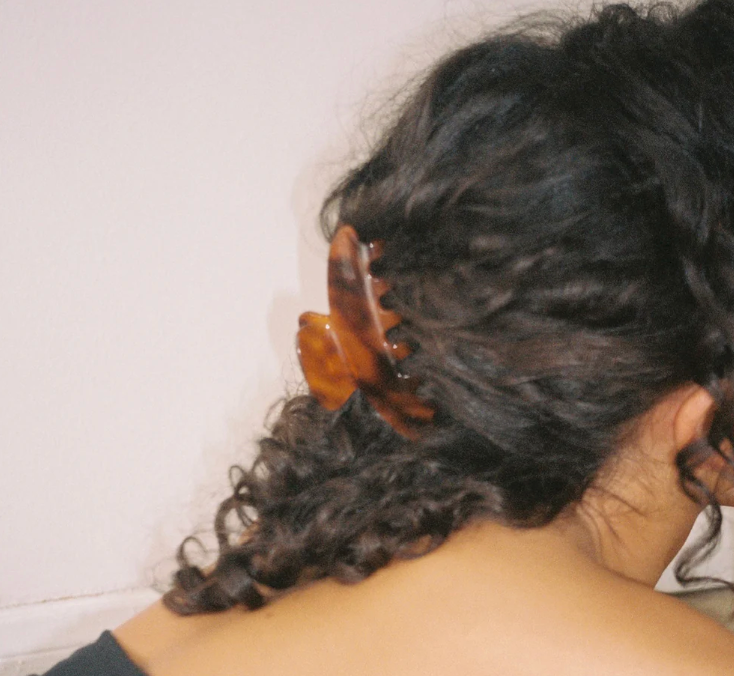 Person with curly hair wearing a wooden hair clip against a white wall