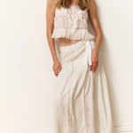 Woman wearing a white ruffled top and long white skirt against a white wall.