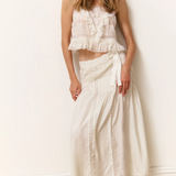 Woman wearing a white ruffled top and long white skirt against a white wall.