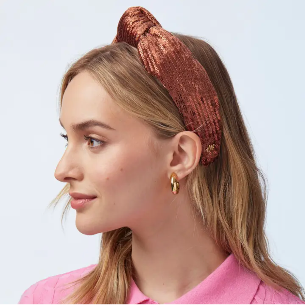Woman wearing a copper sequined knotted headband, gold chunky hoop earrings and a pink top on a white background