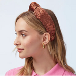Woman wearing a copper sequined knotted headband, gold chunky hoop earrings and a pink top on a white background