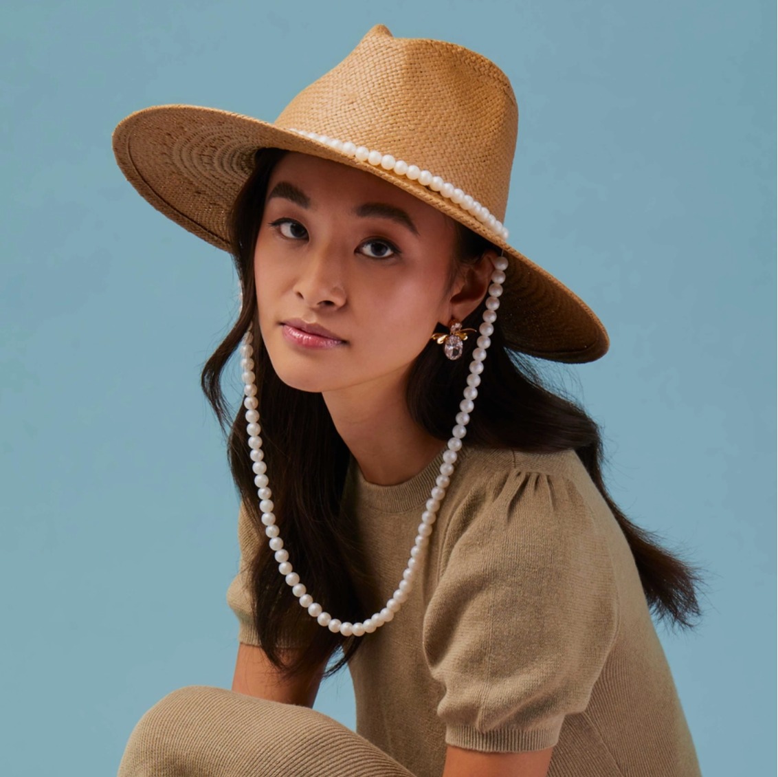 Woman wearing a straw hat with a pearl band and a beige outfit against a blue background