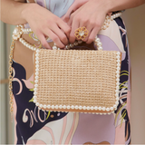 Beige handbag with pearl detailing held by a person wearing a floral dress.