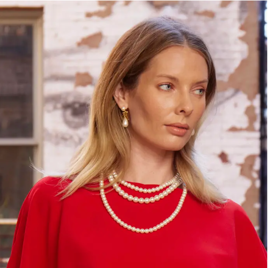 Woman wearing a multi strand pearl necklace and earrings with a red top against a textured wall.