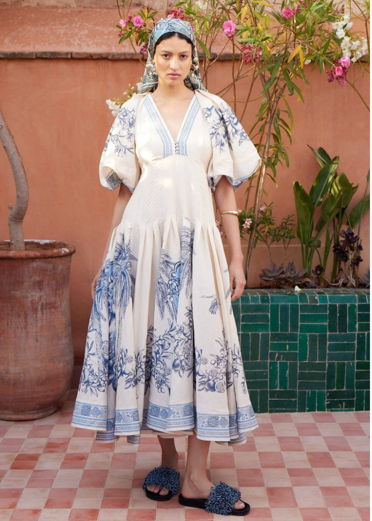 Woman in a white dress with blue floral patterns standing in front of a pink wall with plants.