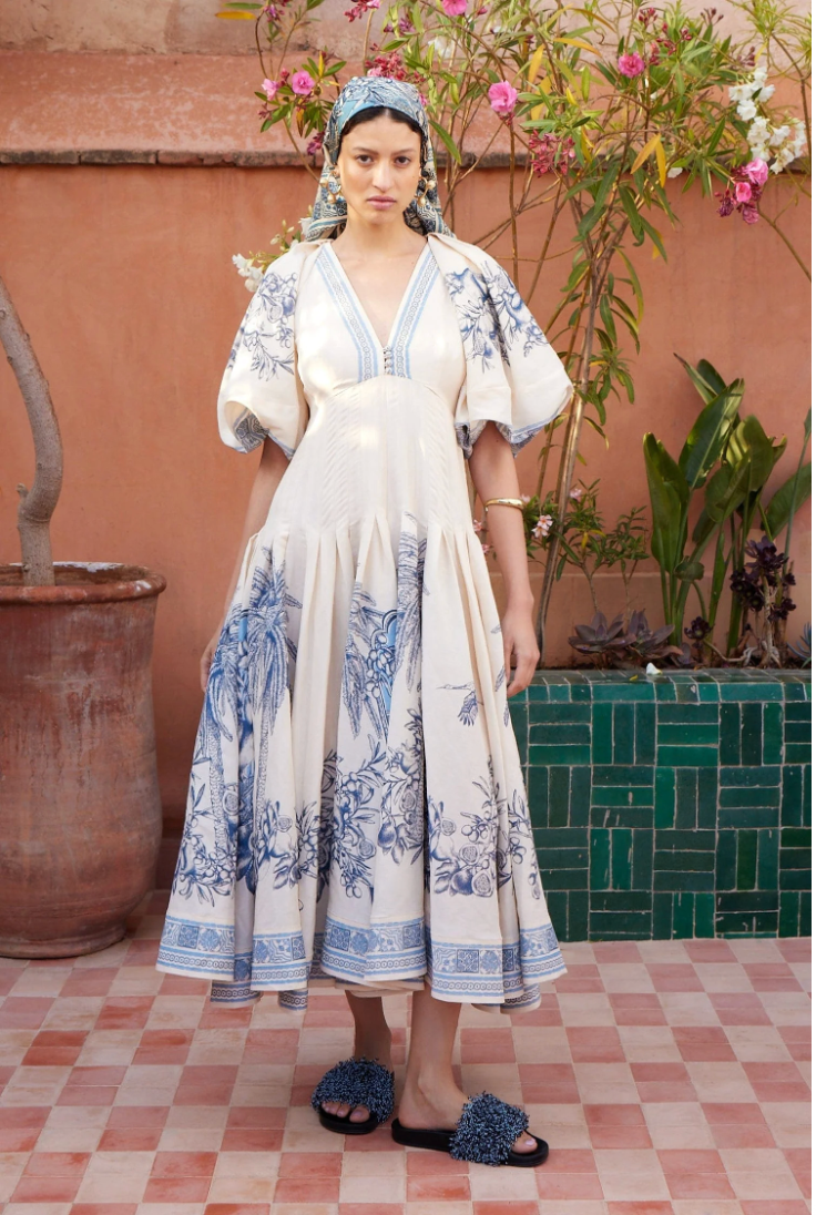 Woman in a white dress with blue floral patterns standing in front of a pink wall with plants.