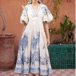Woman in a white dress with blue floral patterns standing in front of a pink wall with plants.