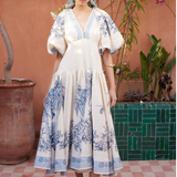 Woman in a white dress with blue floral patterns standing in front of a pink wall with plants.