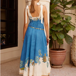 Woman wearing a blue and white dress with floral embroidery, standing in a room with a plant and decorative items.