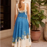 Woman wearing a blue and white dress with floral embroidery, standing in a room with a plant and decorative items.