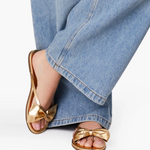 Gold sandals with bow detail worn with blue jeans on a white background