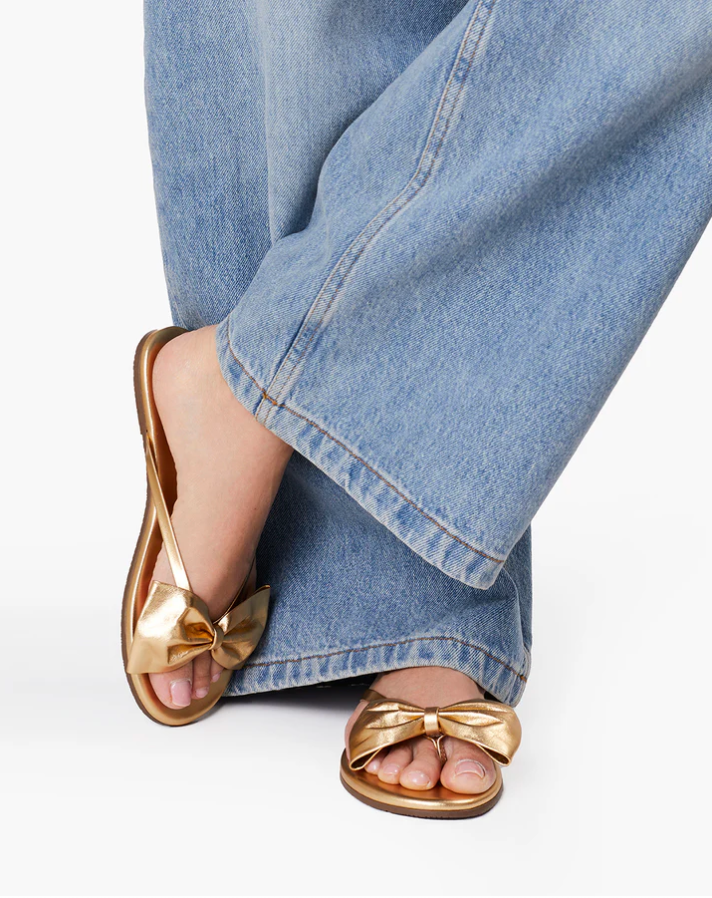 Gold sandals with bow detail worn with blue jeans on a white background