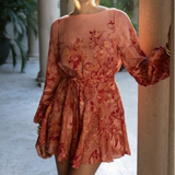 Woman wearing a rust-colored floral dress standing in an outdoor setting with columns and plants.