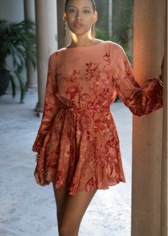Woman wearing a rust-colored floral dress standing in an outdoor setting with columns and plants.