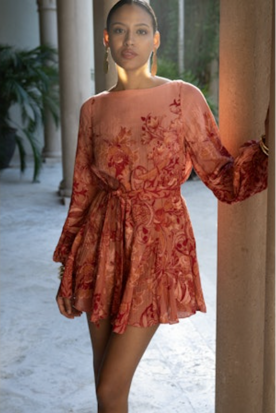 Woman wearing a rust-colored floral dress standing in an outdoor setting with columns and plants.