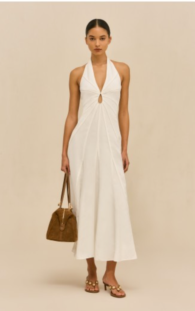 Woman wearing a white halter neck dress holding a brown handbag on a beige background